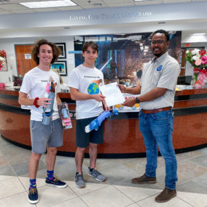Jonathan and David Tamen stand in the lobby at Food For The Poor. They are holding 3D-printed prosthetic hands and forearms. Dwayne Reynolds, Haiti project manager for FFTP, is handing letter of thanks for the donation to David Tamen.