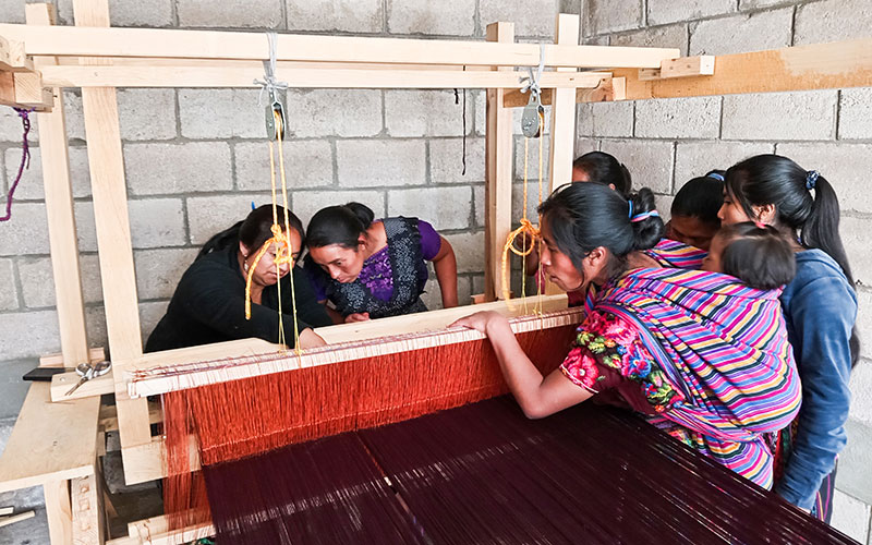 Women engaged in weaving on a machine within a bright room.