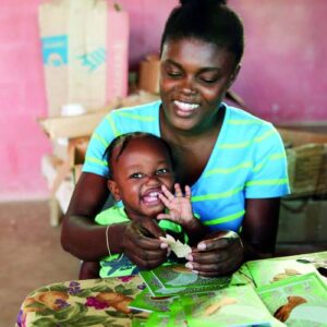 A smiling Haitian mother with a child on her lap makes handcrafted cards for FFTP's gift catalog