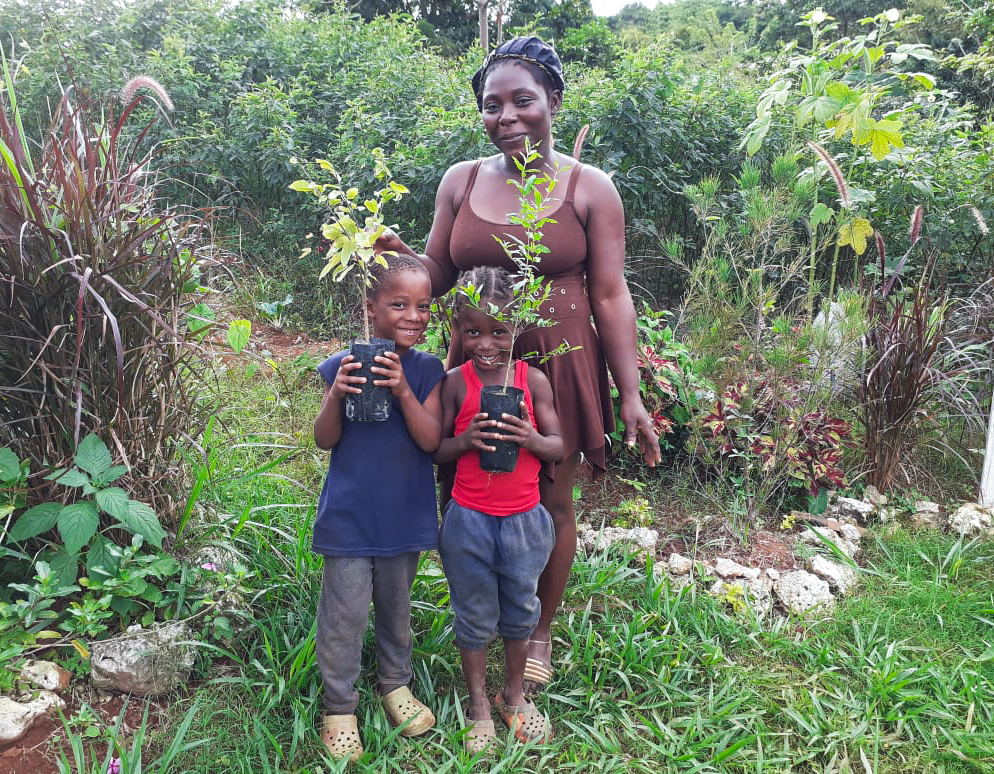 A family in Jamaica. A woman stands behind two children. The children are holding young fruit trees in pots.