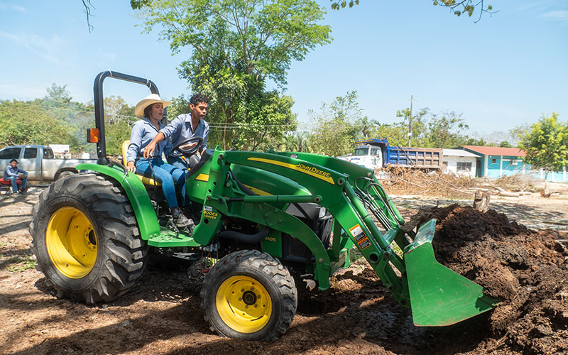A man and woman drive a green tractor through a rural landscape.