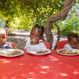 3 children in Haiti sitting at a red table with plates of food thanks to FFTP Giving Tuesday donors