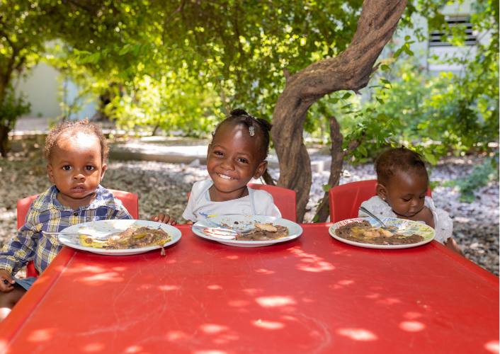 3 children in Haiti sitting at a red table with plates of food thanks to FFTP Giving Tuesday donors