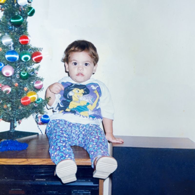 Monica as a toddler, sitting on a bench in front of a Christmas tree