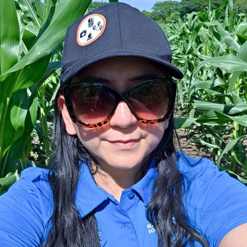 A photo of a woman in a hat and sunglasses smiling in front of a field