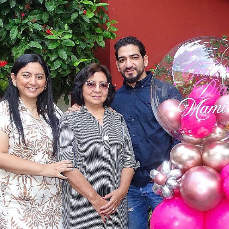 An older woman stands by balloons with Happy Mother's Day written in Spanish, between her daughter and son.