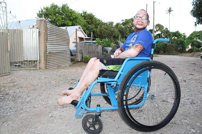 A smiling teenaged boy sits in a sturdy, blue wheelchair with mountain bike tires. The boy, who has vitiligo, is wearing glasses, a blue t-shirt, and black and green basketball shorts.