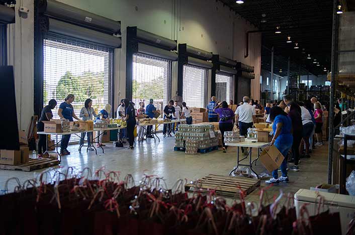 Dozens of volunteers stand at two rows of folding tables in a warehouse. They are packing canned goods and other items into small cardboard boxes. There are pallets of canned goods in between the rows of tables. In the foreground, there is a table of gift bags tied with red ribbon.
