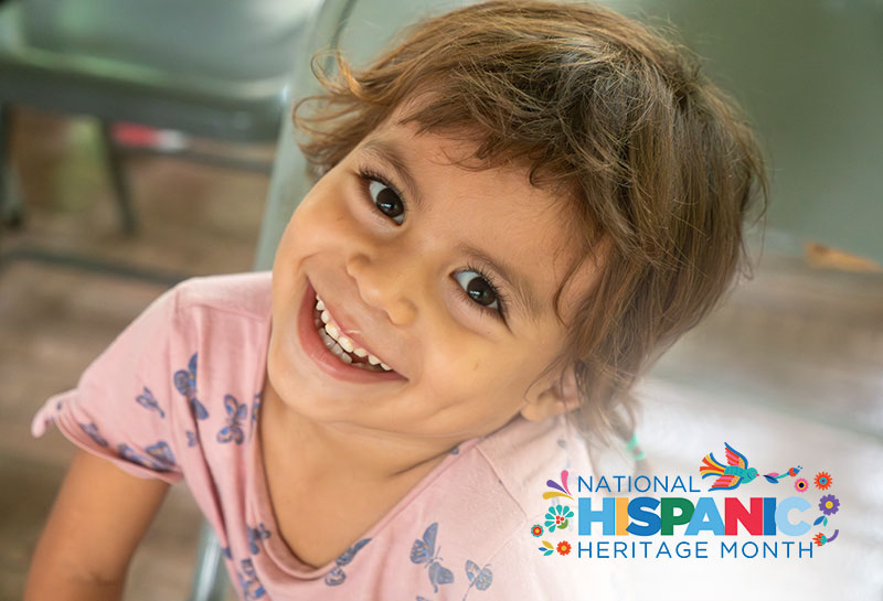 A young girl from Honduras looks at the camera smiling with Hispanic Heritage Month logo