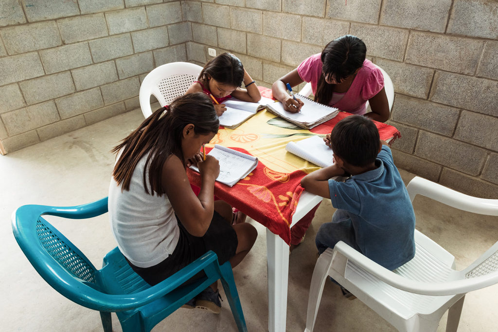 Maria's children sit at a table in their new home, writing in notebooks.