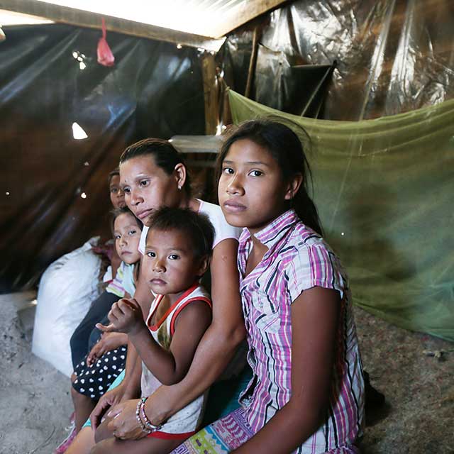Maria and her children sit in their old home made of scraps of wood and plastic tarps.