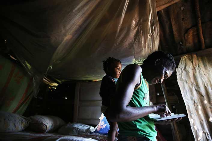 A woman in a makeshift homes reads a Bible. Her young sons stands behind her.