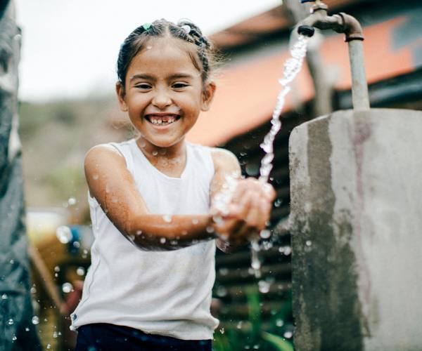 young girl smiles as she puts her hands under an outdoor faucet using safe water to wash her hands
