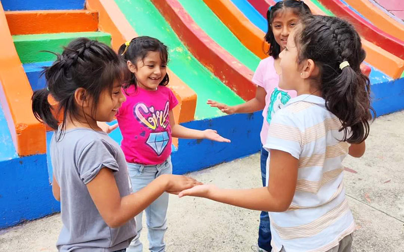 Four girls stand together in front of a vibrant, colorful slide at a playground, smiling. Sponsoring a child through FFTP