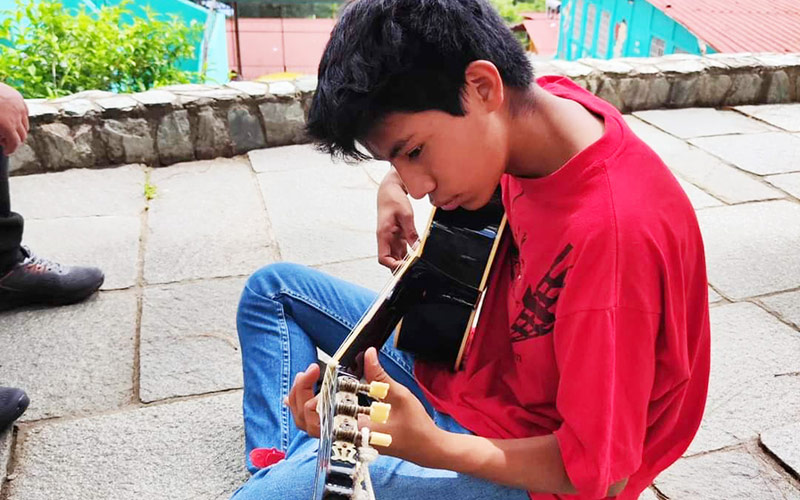 A young man sits on the ground, strumming an acoustic guitar with a focused expression.
