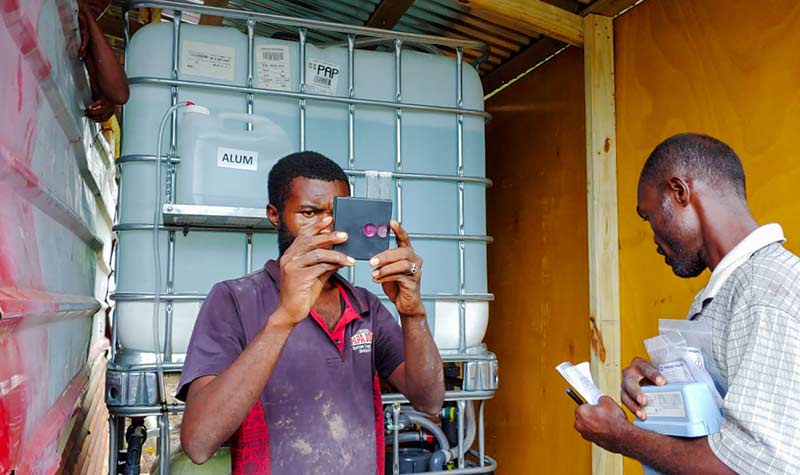 Two men in Haiti stand in front of a water filtration unit that provides treated water.