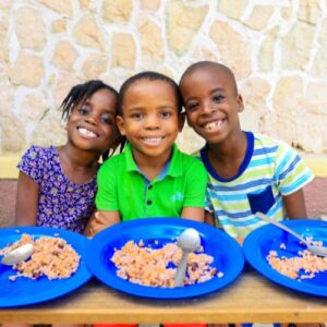 3 Haitian children sit in front of blue plates of food, smiling, triple tuesday