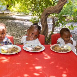 3 young children in Haiti sitting at a red table with plates of food