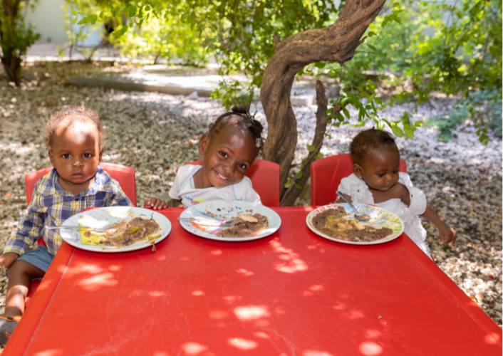 3 young children in Haiti sitting at a red table with plates of food