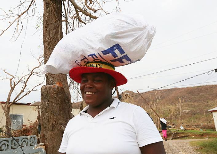 A person in Jamaica carrying a large relief kit on their head, smiling, united for relief concert