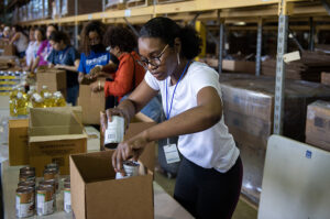 A volunteer in a warehouse packs canned goods into a cardboard box. She is wearing a lanyard with a badge that says "Volunteer." A row of volunteers is visible in the background.