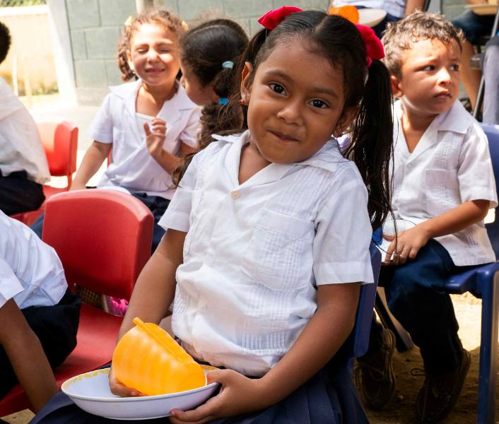 A young girl in Honduras sits in a feeding center with other children holding a plate and cup and smiling