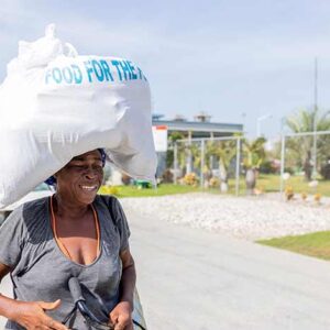 A woman walks down a street in Haiti carrying a large white bag on her head.