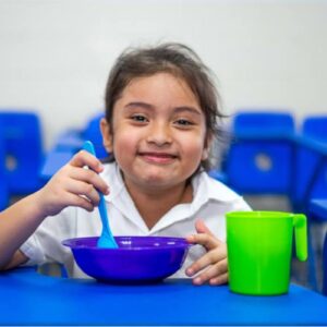 A young girl sitting at a blue table eating from a blue bowl, smiling